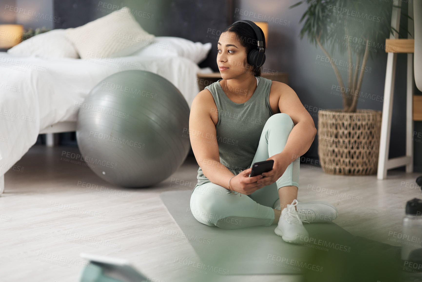 Buy stock photo Shot of a sporty young woman wearing headphones and using a cellphone while exercising at home