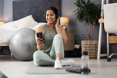 Buy stock photo Shot of a sporty young woman wearing headphones and using a cellphone while exercising at home