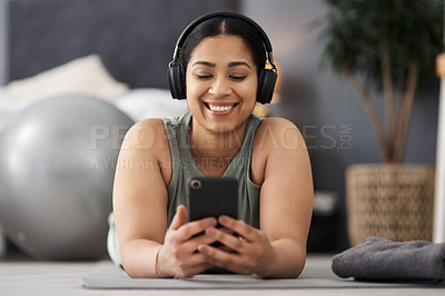 Buy stock photo Shot of a sporty young woman wearing headphones and using a cellphone while exercising at home