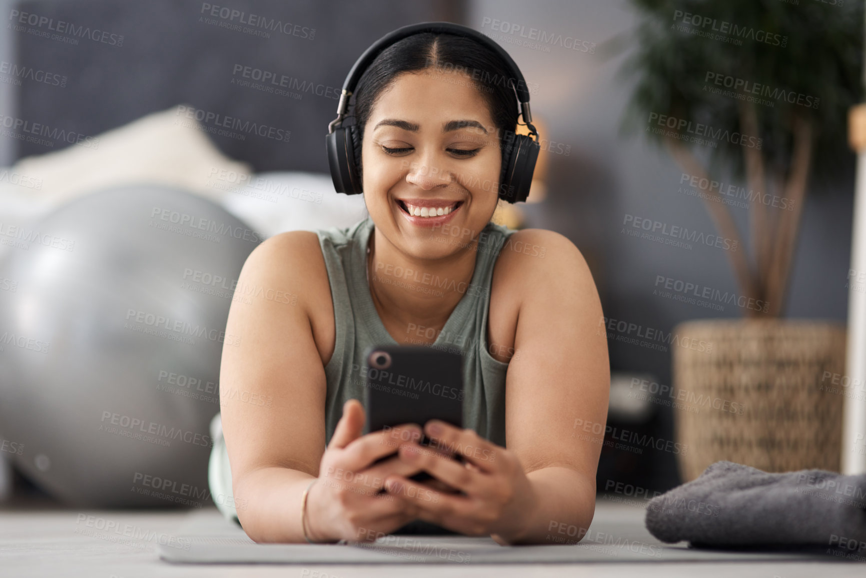 Buy stock photo Shot of a sporty young woman wearing headphones and using a cellphone while exercising at home