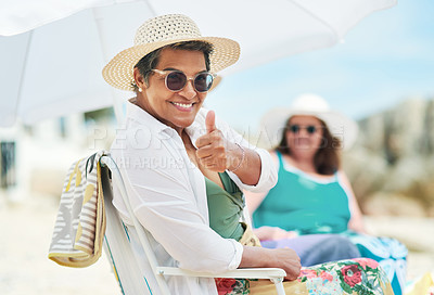 Buy stock photo Shot of a mature woman sitting and making a thumbs up gesture during a day out on the beach with friends