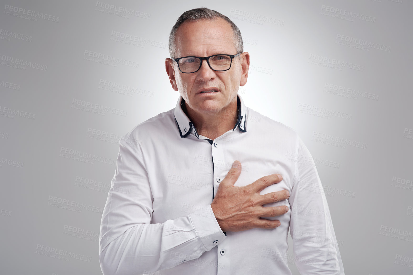 Buy stock photo Shot of a handsome mature businessman standing alone against a grey background in the studio and clutching his chest