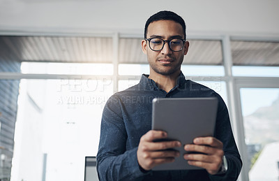 Buy stock photo Shot of a handsome young businessman standing alone in his office and using a digital tablet