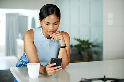 Buy stock photo Shot of an attractive young woman leaning on her kitchen counter and looking contemplative while using her cellphone