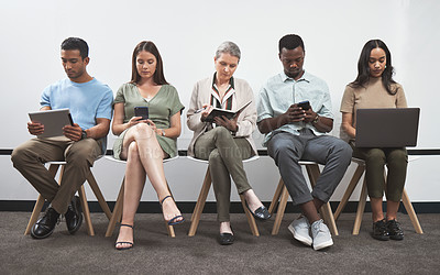 Buy stock photo Shot of a group of businesspeople using digital devices while sitting together in a line against a white wall
