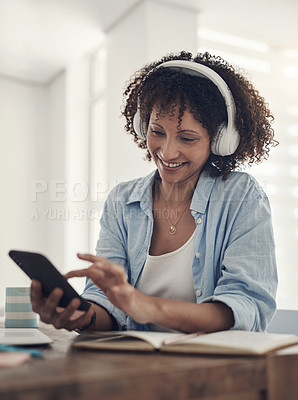 Buy stock photo Shot of an attractive young woman sitting alone at home and using her cellphone while wearing headphones