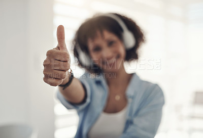 Buy stock photo Defocused shot of a young woman sitting alone at home and showing a thumbs up gesture while wearing headphones