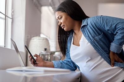 Buy stock photo Shot of a pregnant woman using her cellphone while sitting at home with her laptop