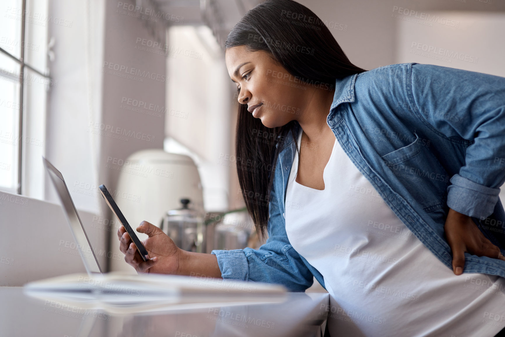 Buy stock photo Shot of a pregnant woman using her cellphone while sitting at home with her laptop