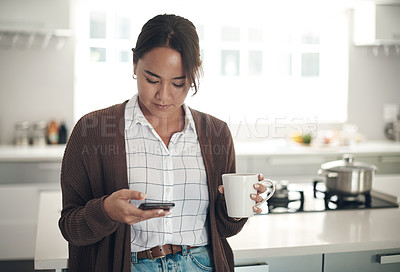 Buy stock photo Shot of a young woman using a cellphone while drinking coffee in the kitchen at home