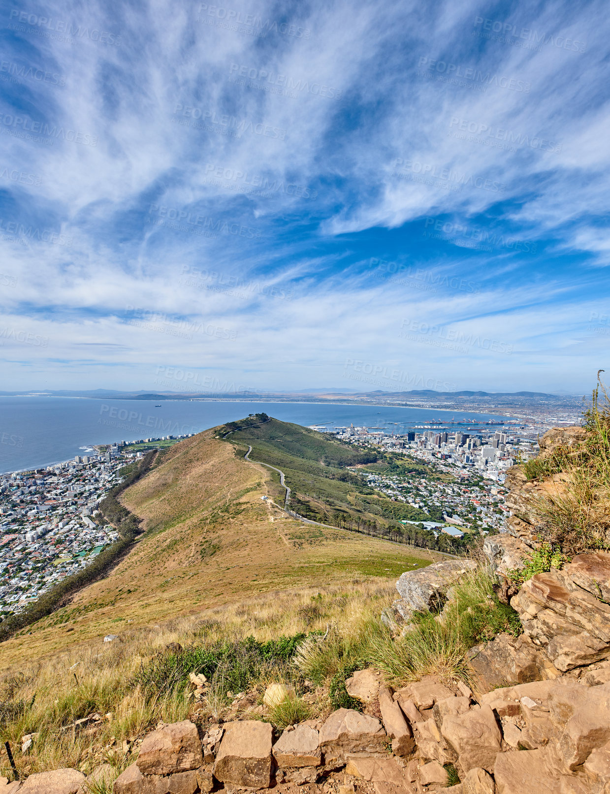 Buy stock photo Rocky mountain with view of a costal city by the sea in Cape Town. Landscape of green hill top with stones and plants surrounded by an urban city and the ocean on a cloudy blue sky with copy space. 