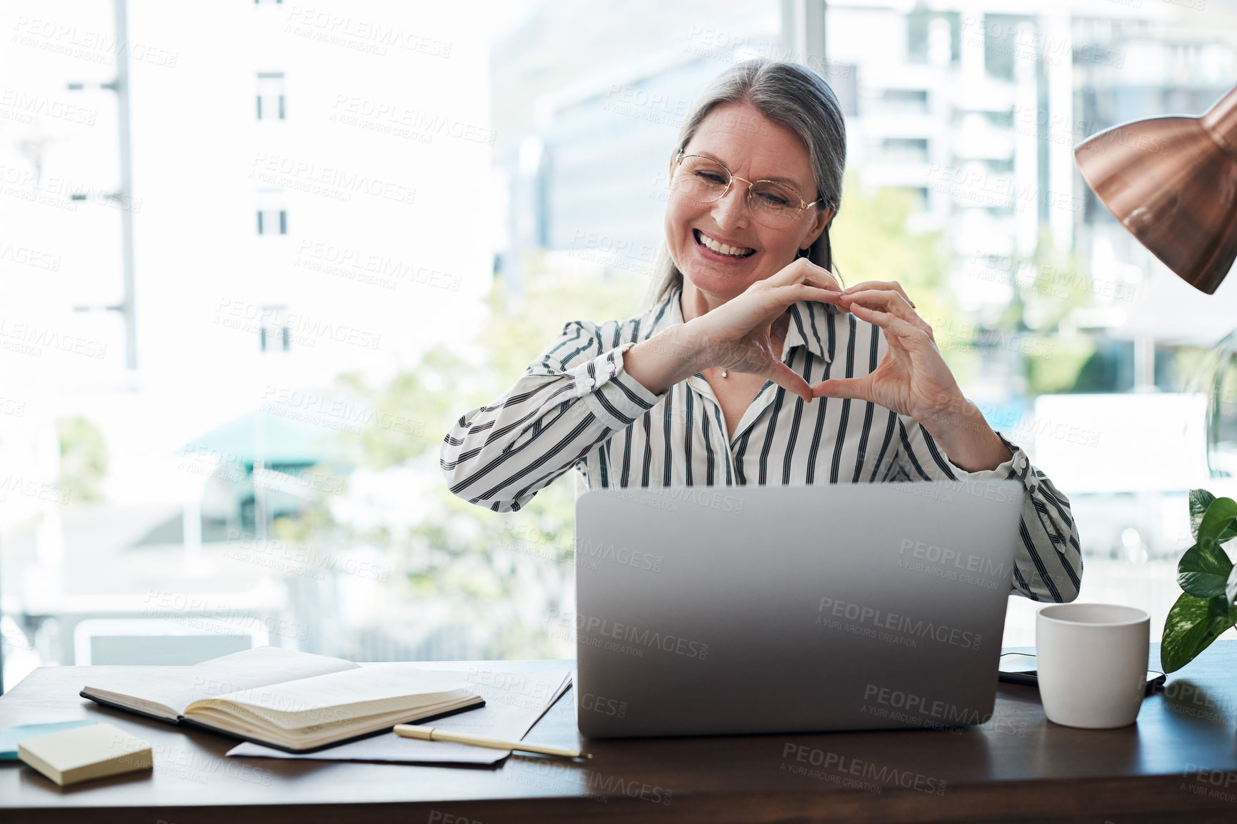 Buy stock photo Shot of a mature businesswoman with a heart formed hand gesture video chatting on her laptop in a modern office