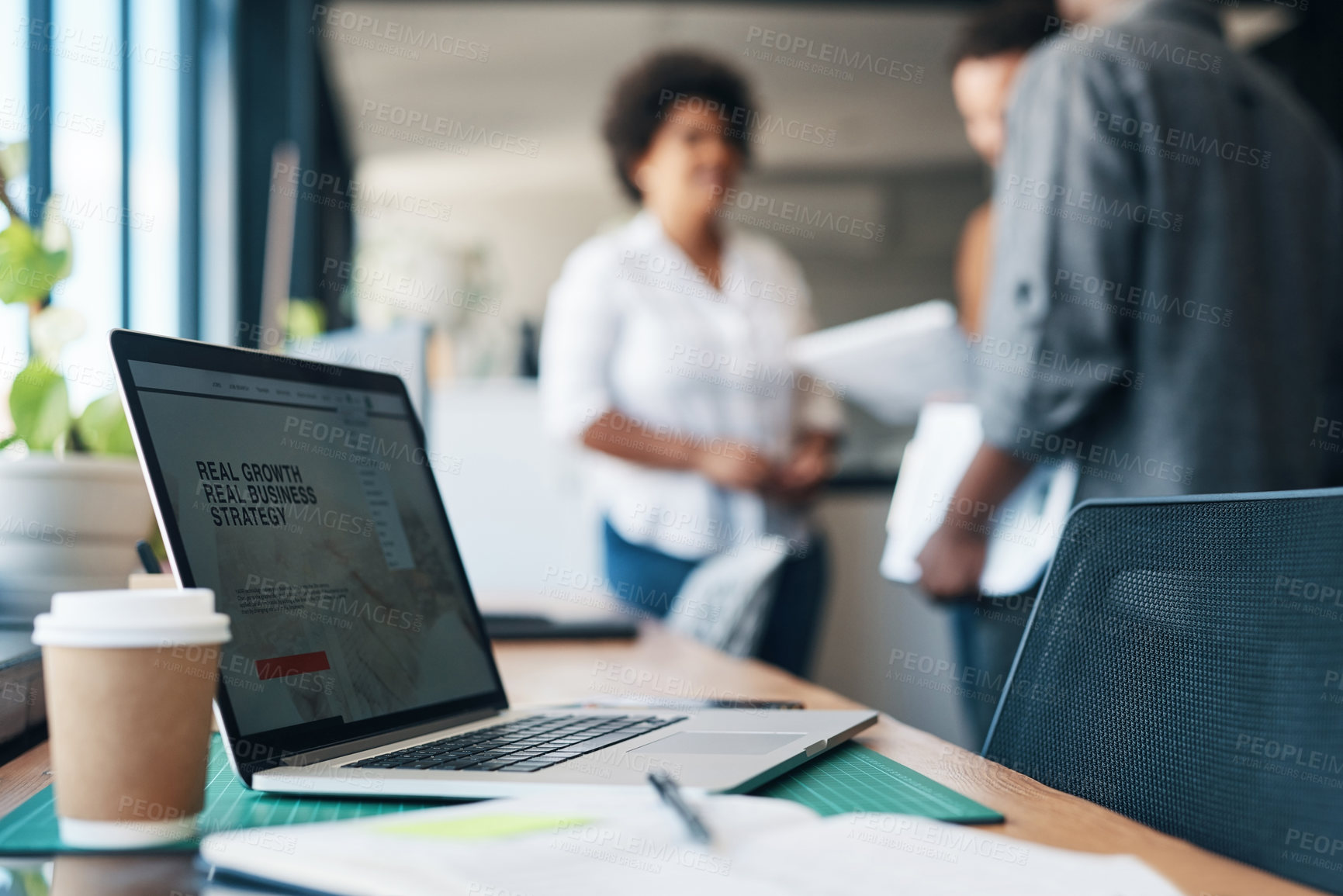 Buy stock photo Shot of a cup of coffee, a notebook and a laptop with a group of businesspeople in the background