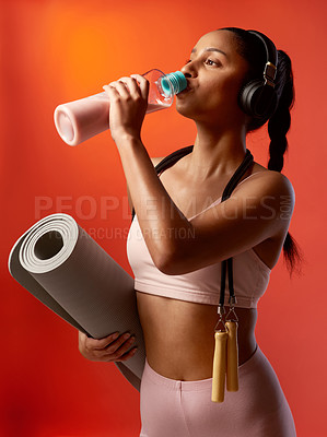 Buy stock photo Studio shot of a sporty young woman drinking water and holding a yoga mat against a red background