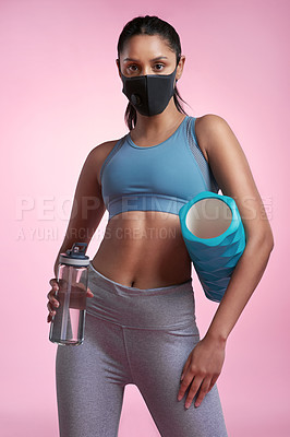 Buy stock photo Cropped portrait of an attractive and sporty young woman wearing a mask while posing with a foam roller and water bottle in studio against a pink background