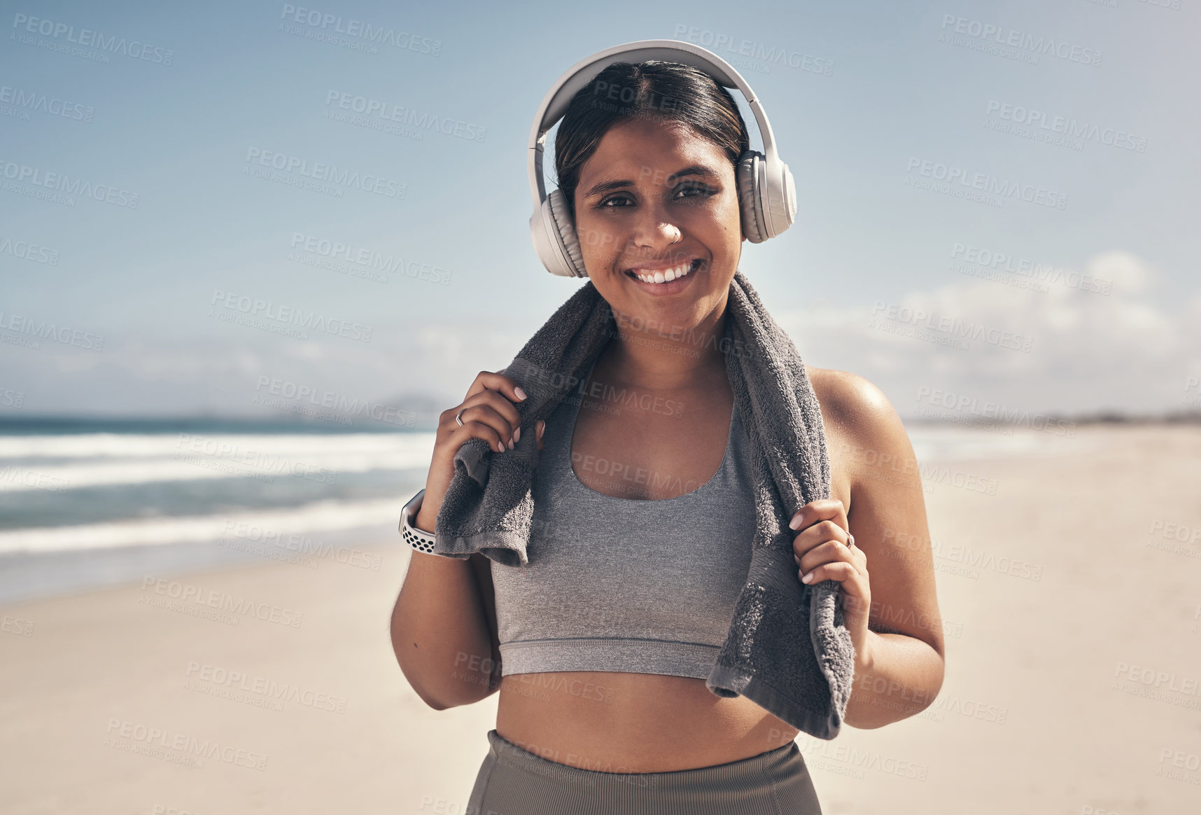 Buy stock photo Shot of a sporty young woman wearing headphones and a towel around her neck on the beach