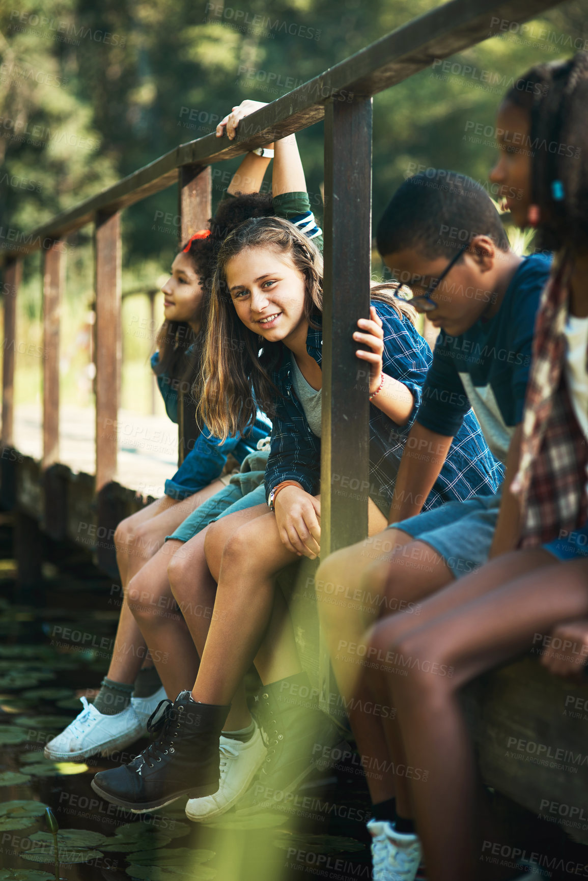 Buy stock photo Shot of a group of teenagers sitting on a bridge in nature at summer camp