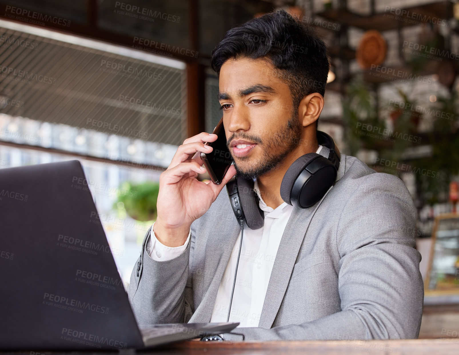 Buy stock photo Shot of a young businessman using a laptop while on the phone at a cafe