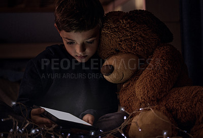 Buy stock photo Shot of an adorable little boy using a digital tablet with his teddy bear at night in his bedroom