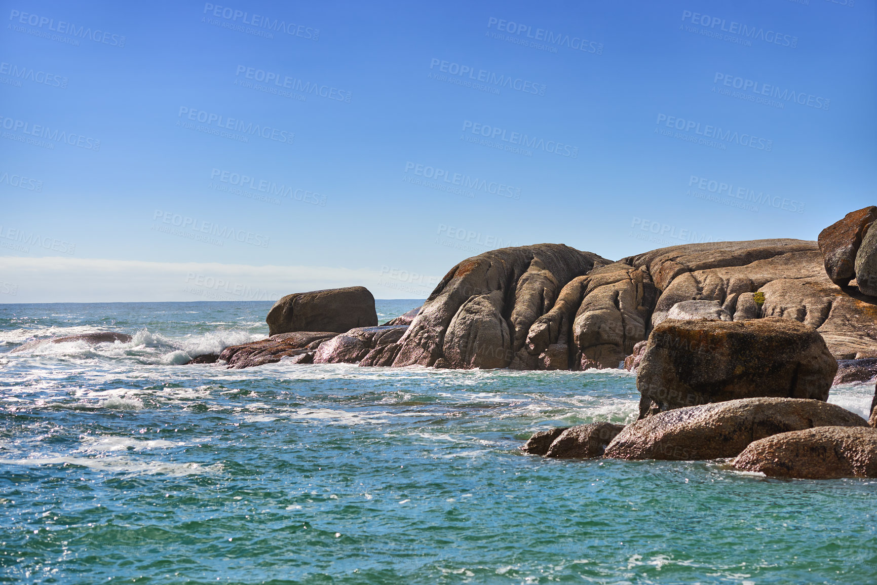 Buy stock photo Scenic view of nature, rocks and calm ocean water on a sunny day in Summer, with a beautiful blue sky in the background. Sea waves breaking close to the shore of the beach.