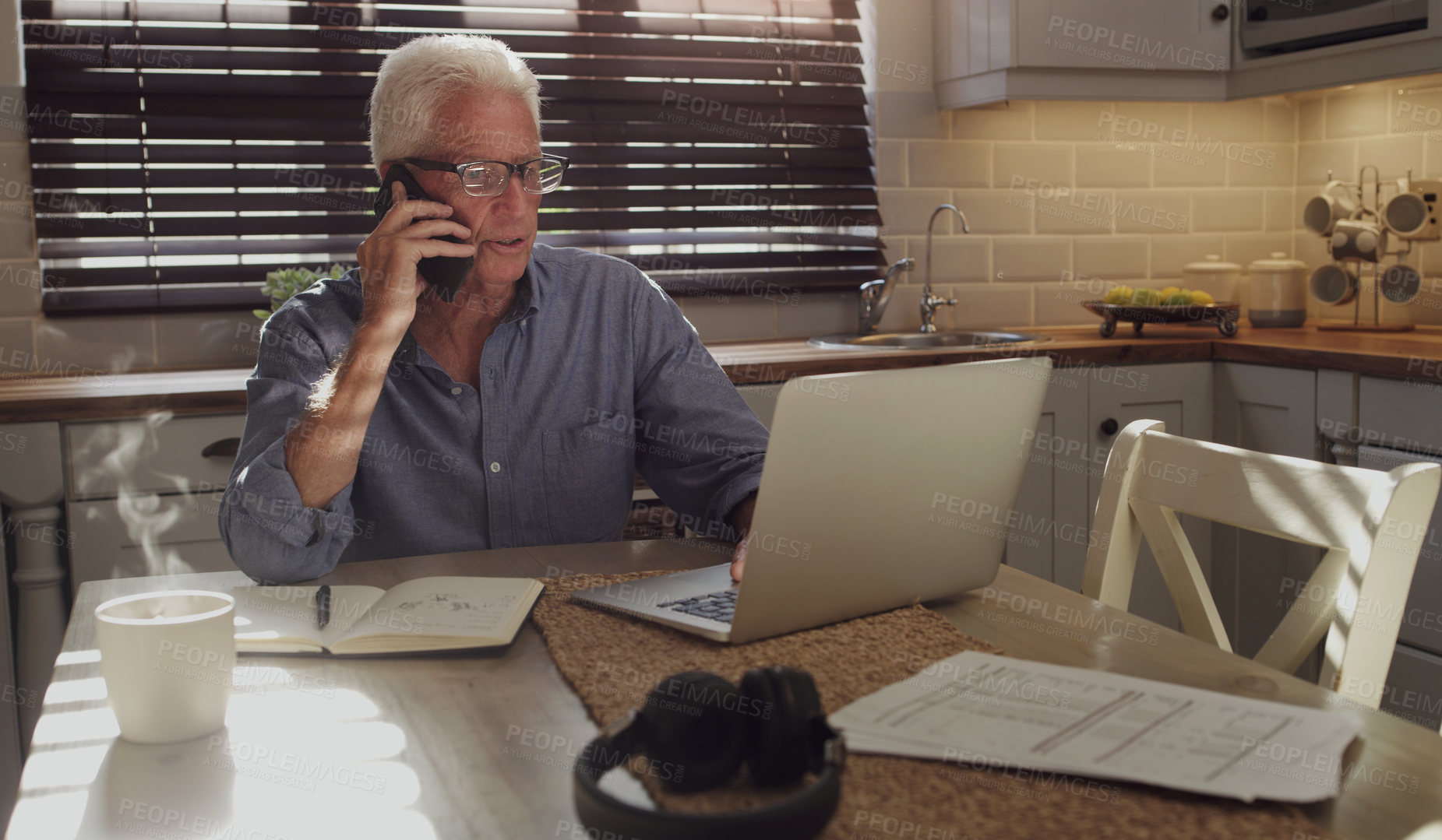 Buy stock photo Shot of a senior man sitting alone in the kitchen and using technology to work from home