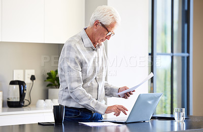 Buy stock photo Shot of an elderly man using his laptop while reading some paperwork
