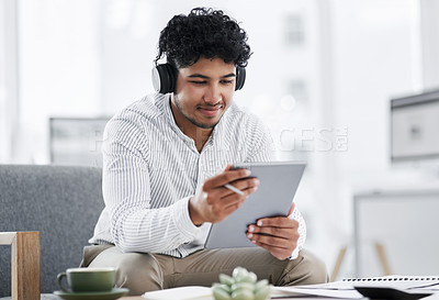 Buy stock photo Shot of a young businessman wearing headphones while using a digital tablet in an office