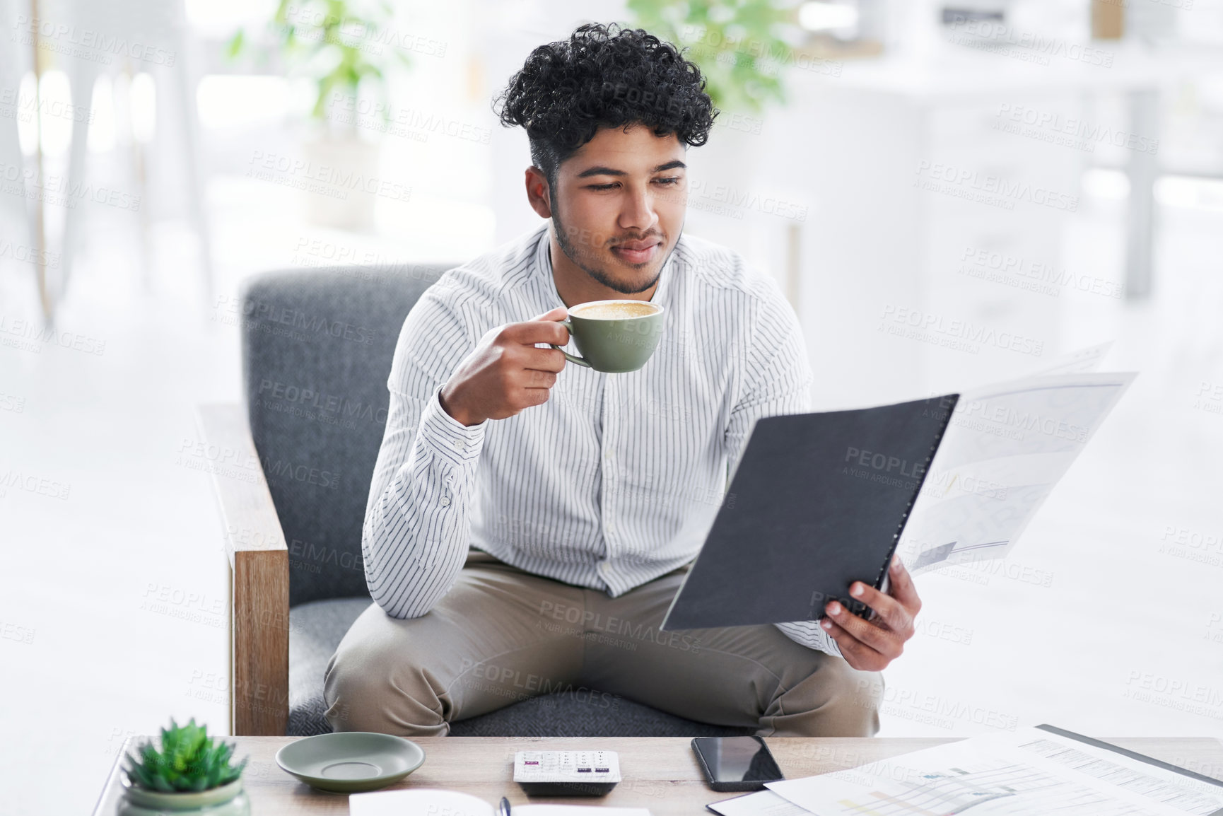 Buy stock photo Shot of a young businessman drinking coffee while going through paperwork in an office