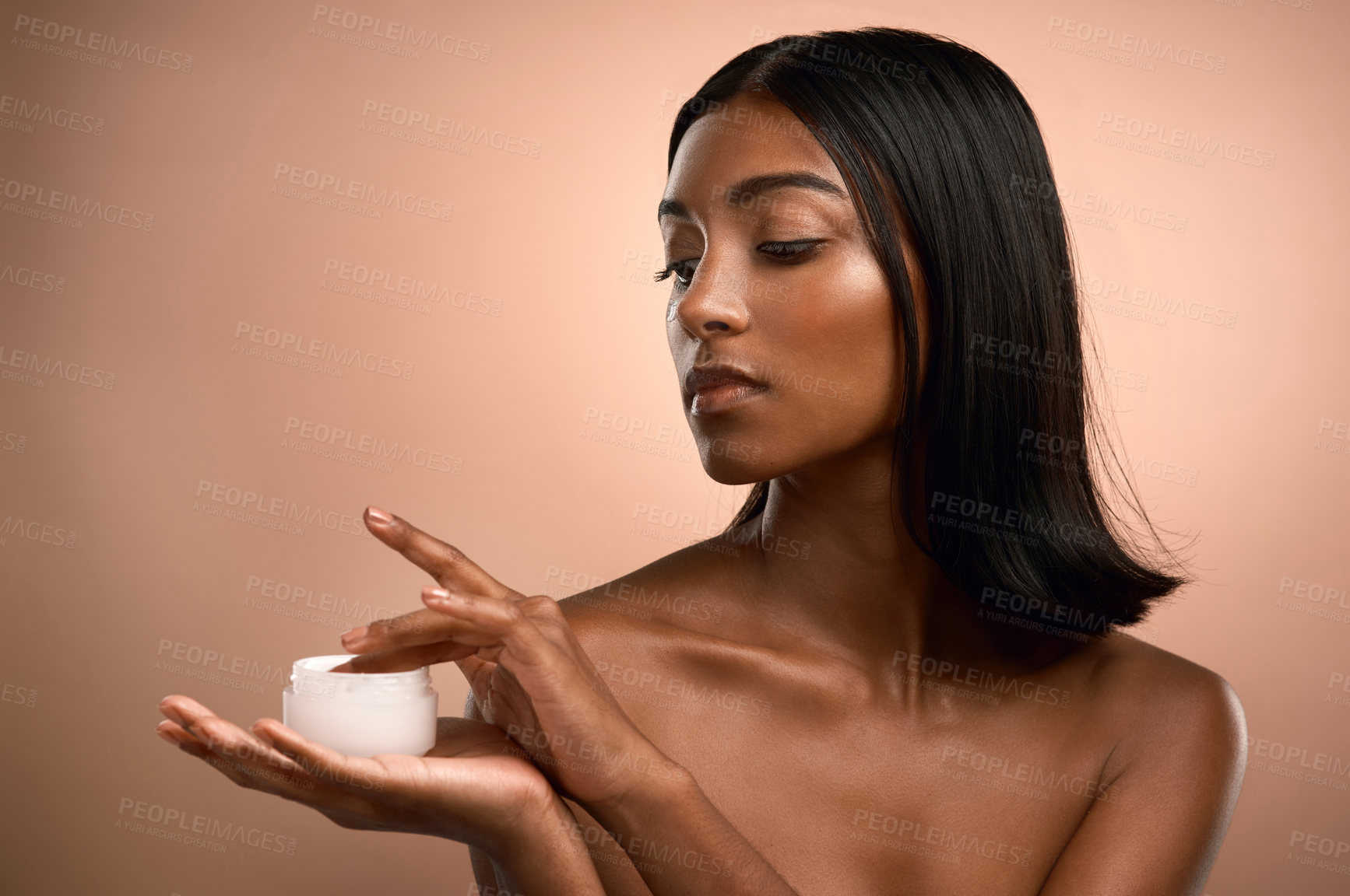 Buy stock photo Shot of an attractive young woman applying moisturiser against a brown background