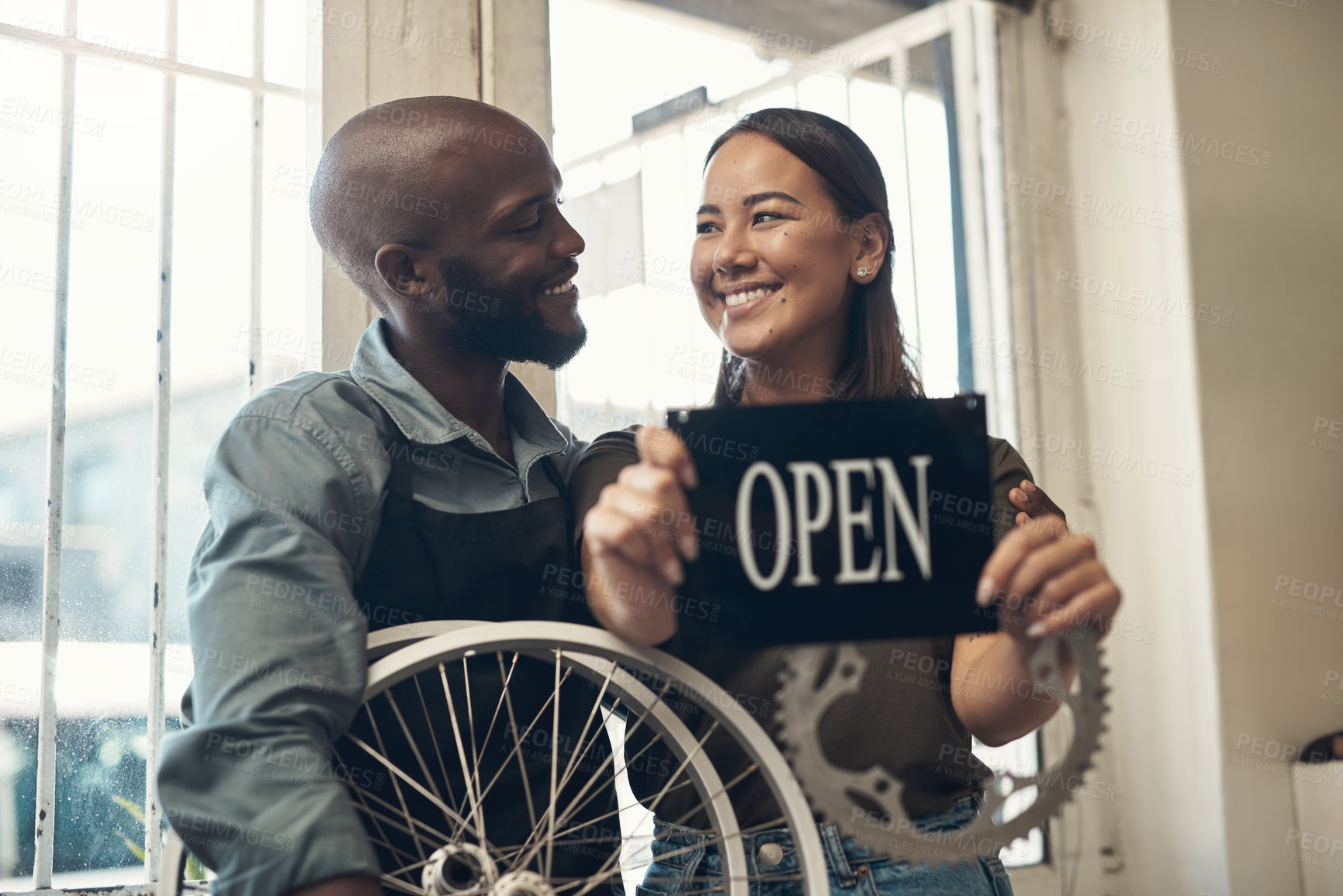 Buy stock photo Shot of two young business owners standing together in their bicycle shop and holding an open sign