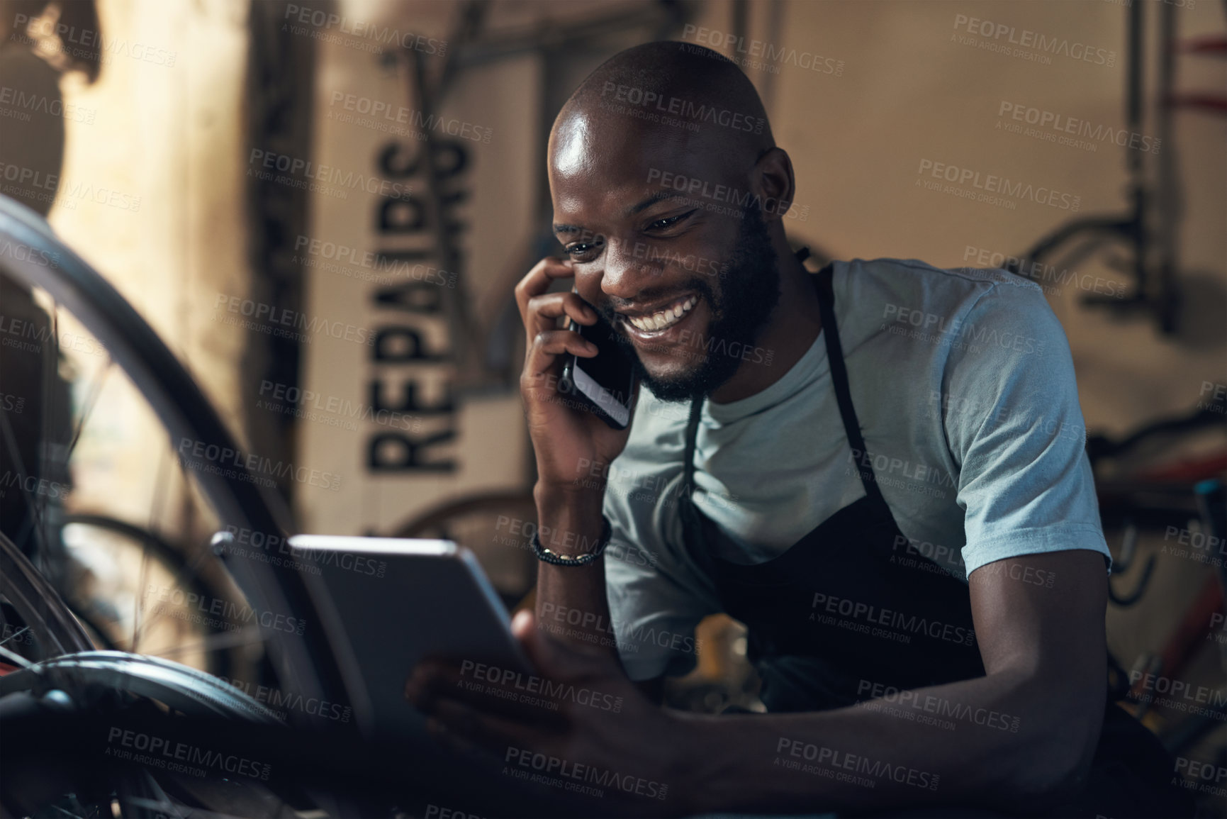Buy stock photo Shot of a young man talking on a cellphone and holding a digital tablet while working at a bicycle repair shop