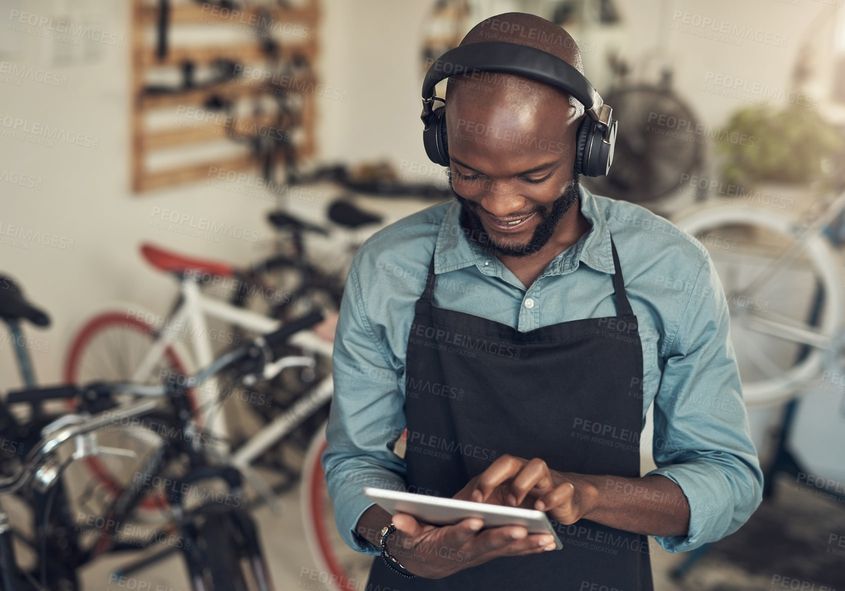 Buy stock photo Shot of a handsome young man standing alone in his bicycle shop and using a digital tablet while wearing headphones