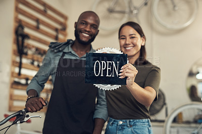 Buy stock photo Shot of two young business owners standing together in their bicycle shop and holding an open sign
