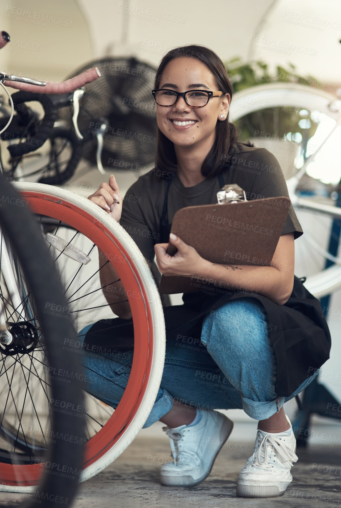 Buy stock photo Full length shot of an attractive young woman crouching in her bicycle shop and holding a clipboard