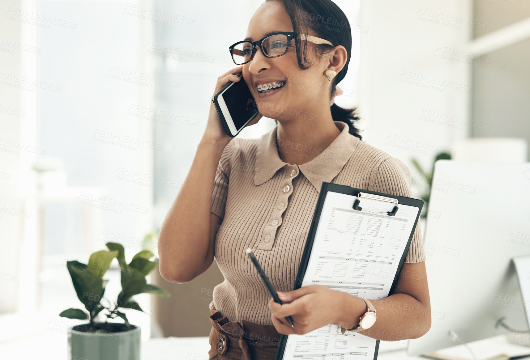 Buy stock photo Shot of a young businesswoman talking on a cellphone while holding a clipboard in an office