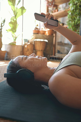 Buy stock photo Shot of a young woman wearing headphones and using a cellphone while lying on an exercise mat at home