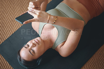 Buy stock photo High angle shot of a young woman wearing headphones and using a cellphone while lying on an exercise mat at home