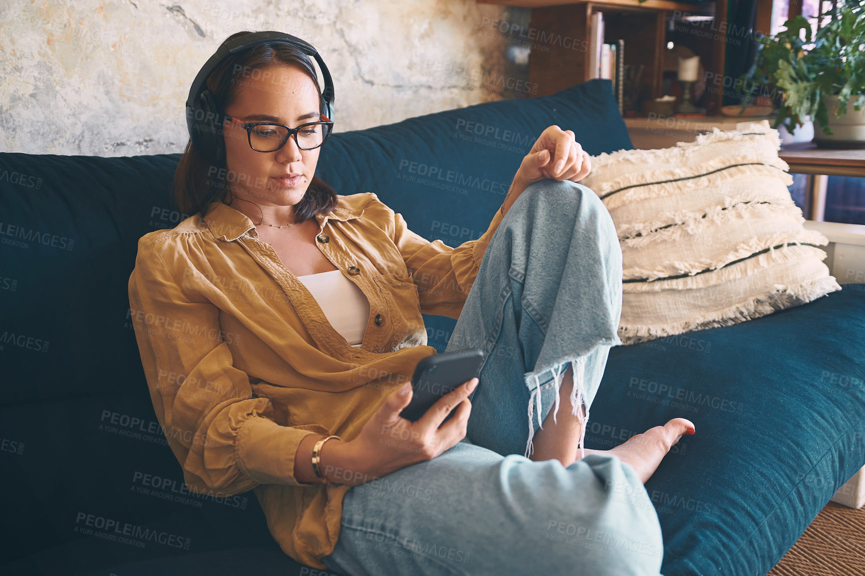 Buy stock photo Shot of a young woman using headphones and a smartphone while relaxing on the sofa at home