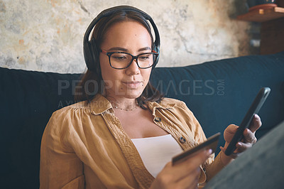 Buy stock photo Shot of a young woman using a smartphone and credit card and headphones on the sofa at home