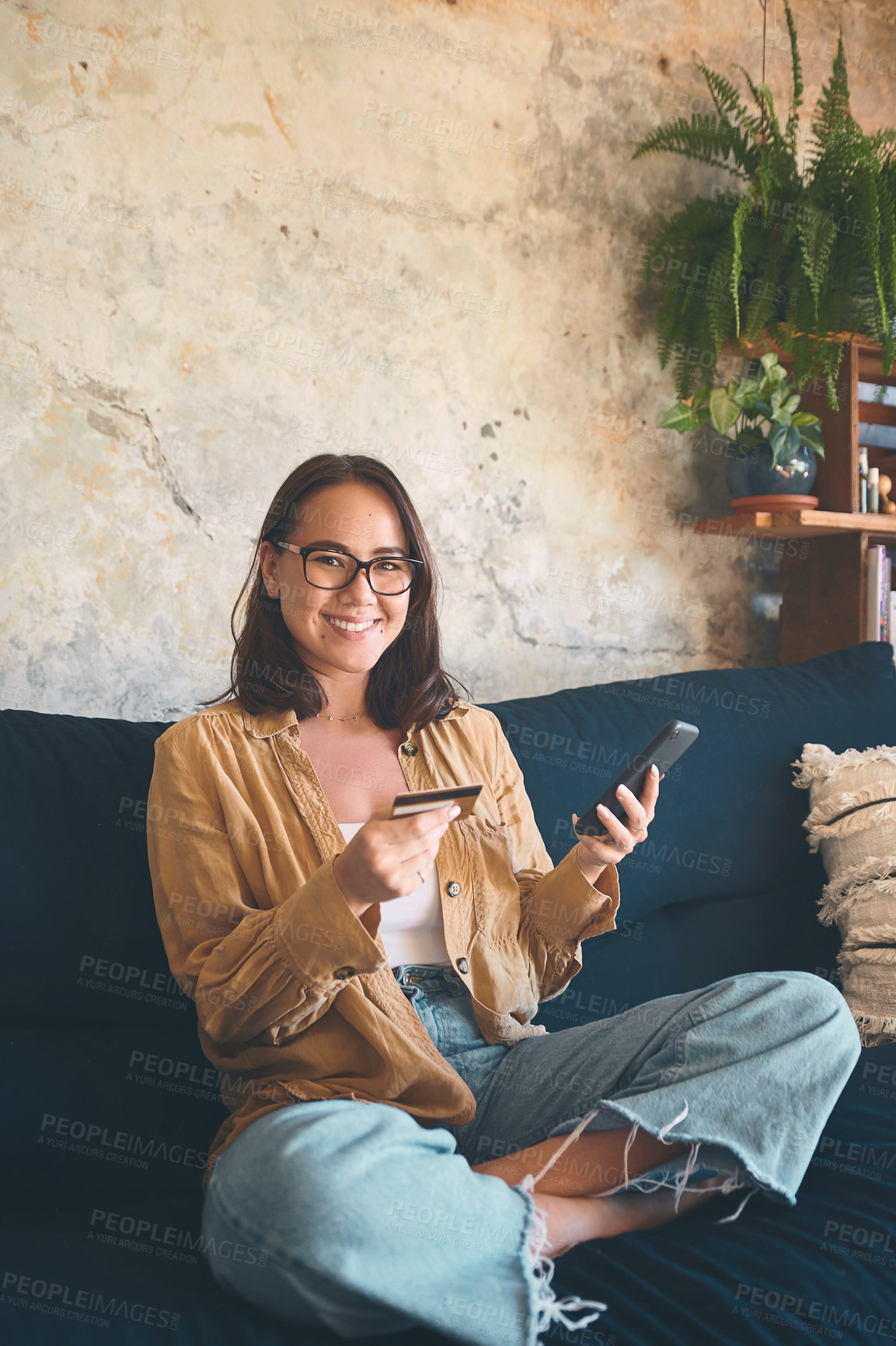 Buy stock photo Shot of a young woman using a smartphone and credit card on the sofa at home