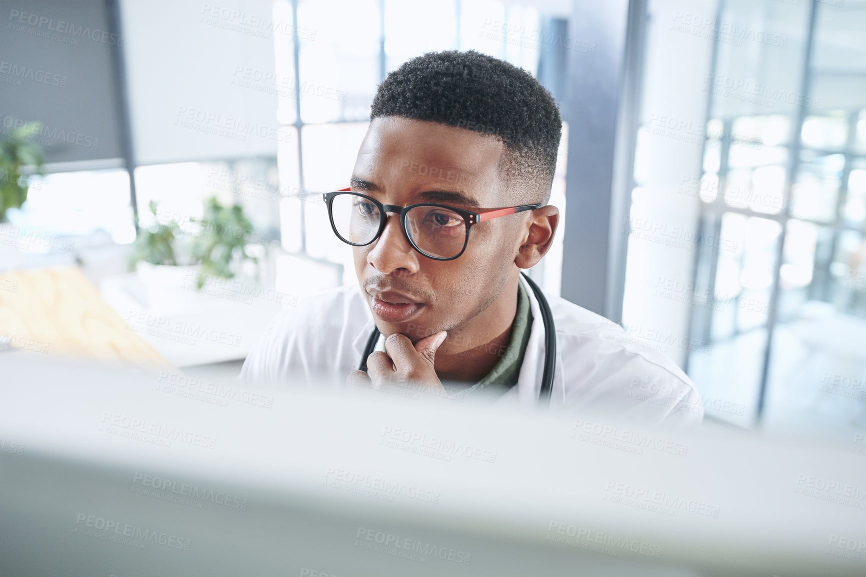 Buy stock photo Shot of a young doctor sitting alone in his office at the clinic and looking contemplative while using his computer
