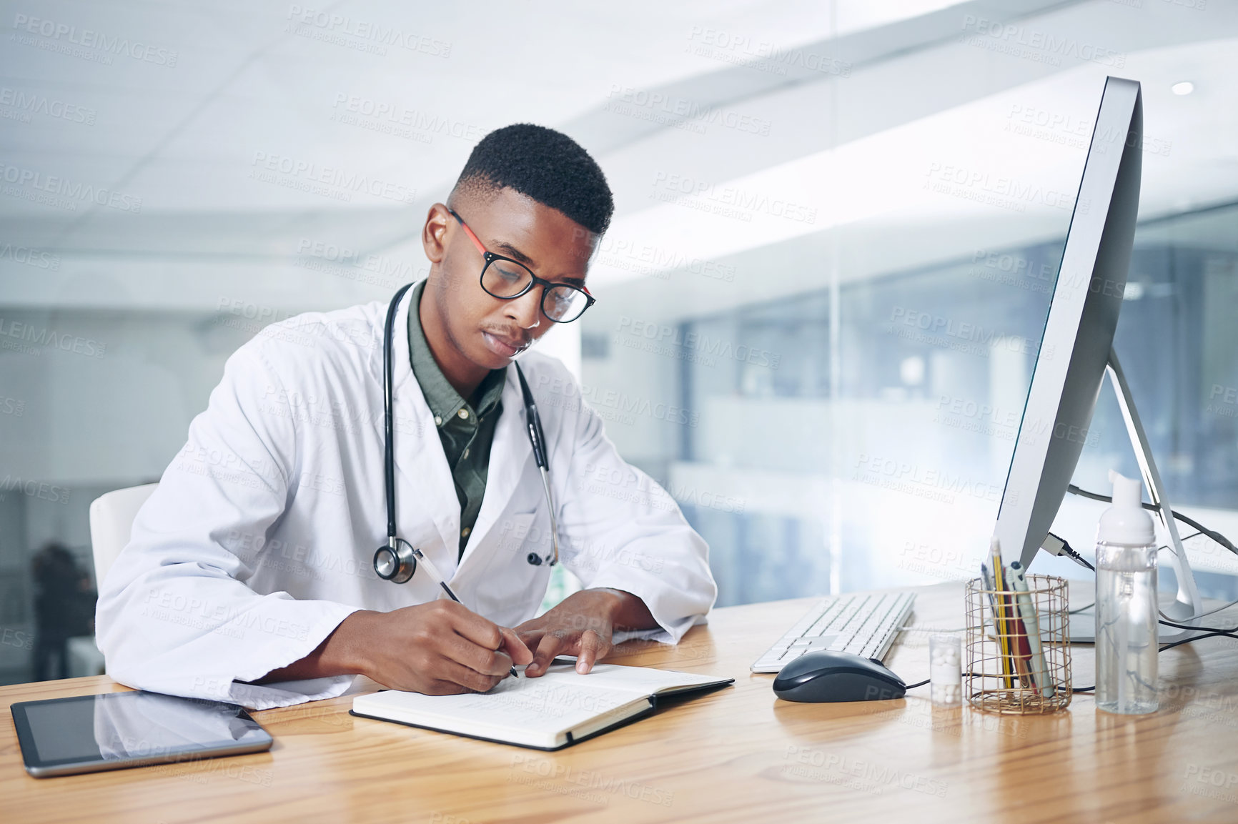 Buy stock photo Shot of a handsome young doctor sitting alone in his office at the clinic and writing in his notebook