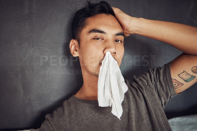 Buy stock photo Shot of a young man blowing his nose while recovering from an illness in bed at home