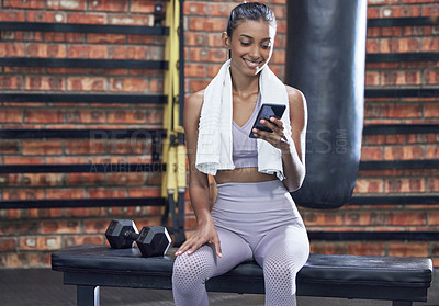 Buy stock photo Shot of a sporty young woman using a cellphone while taking a break in a gym