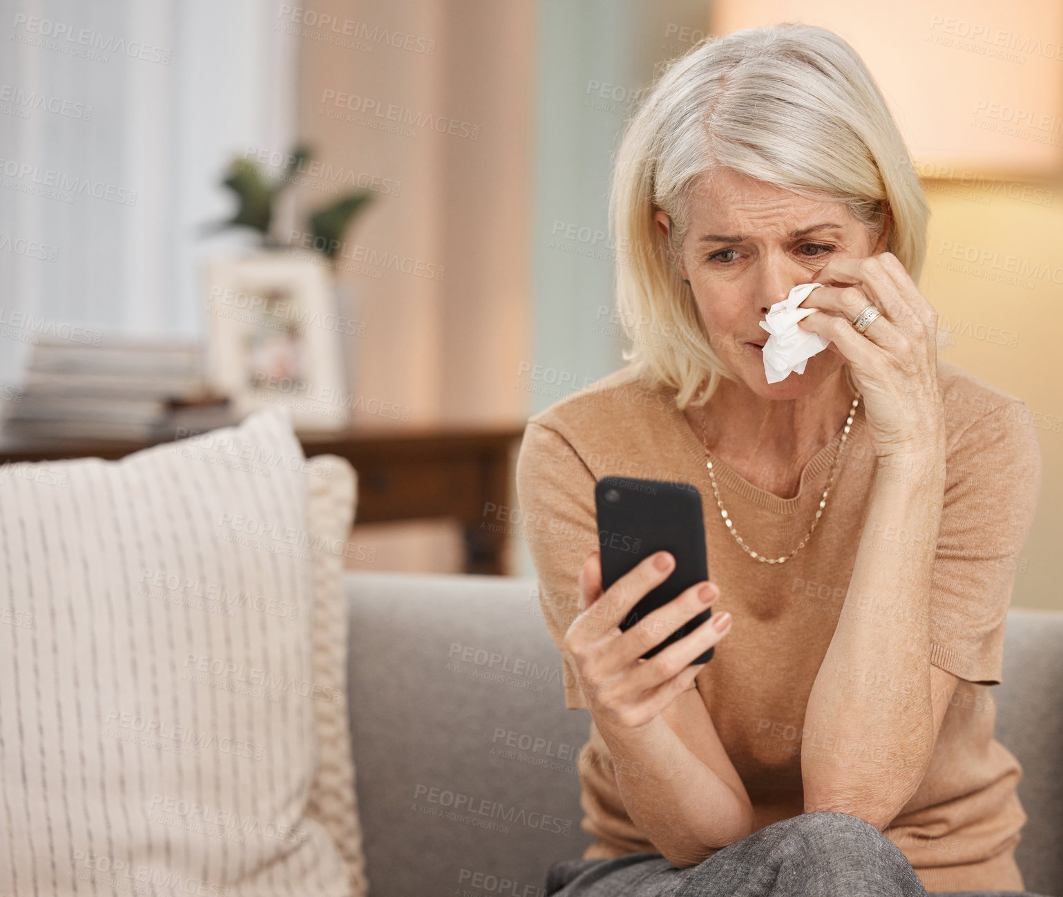 Buy stock photo Shot of a mature woman using a smartphone and crying on the sofa at home