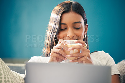 Buy stock photo Shot of a young woman sitting in her living room and enjoying a cup of tea while using her laptop