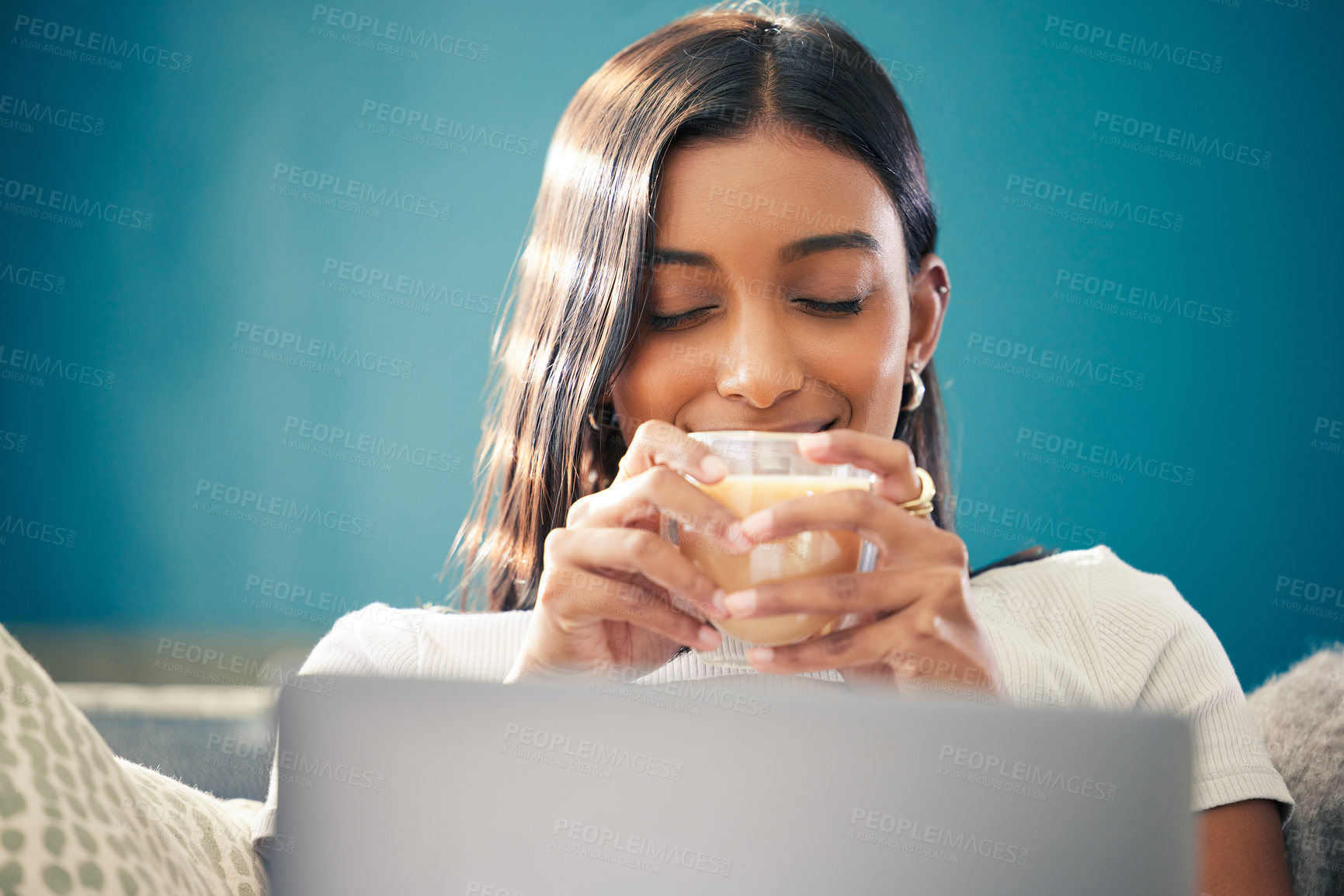 Buy stock photo Shot of a young woman sitting in her living room and enjoying a cup of tea while using her laptop