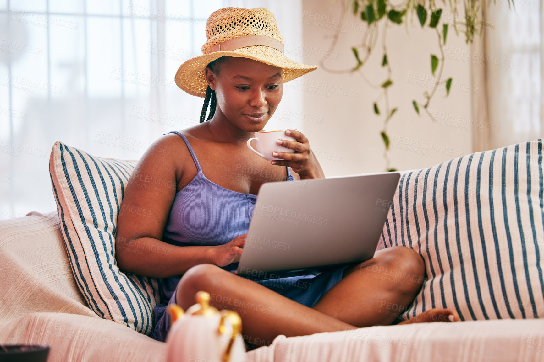 Buy stock photo Shot of a young woman wearing a sunhat and using her laptop while having a coffee break on the sofa