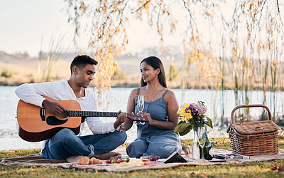 Buy stock photo Shot of a young man playing a guitar while on a picnic with his girlfriend at a lakeside