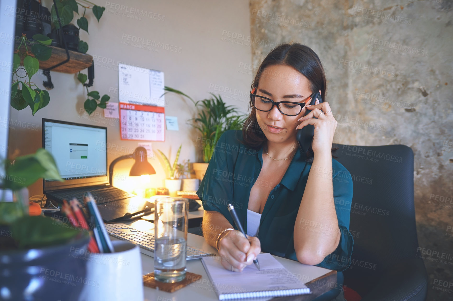 Buy stock photo Shot of an attractive young woman sitting alone and using her cellphone while writing notes in her home office